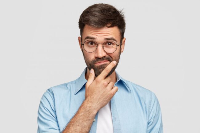 Image of hesitant unshaven European male with thick beard, holds chin, purses lips with clueless expressions, doubts what to eat delicious for supper, isolated over white background. What to choose? cómo sé si tengo bruxismo