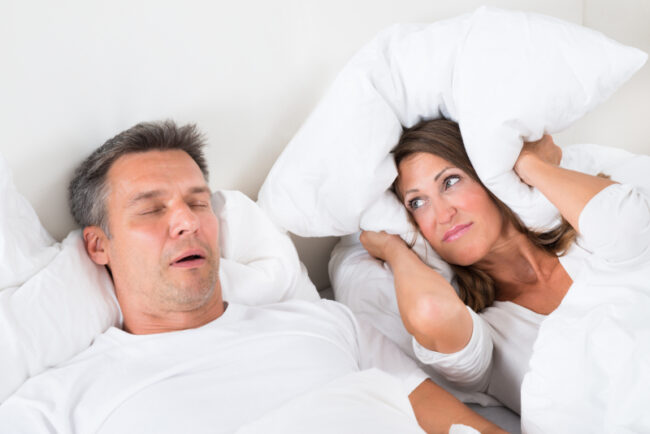 Woman,Covering,Her,Ears,With,Pillow,While,Man,Snoring tratamiento de la apnea del sueño en madrid